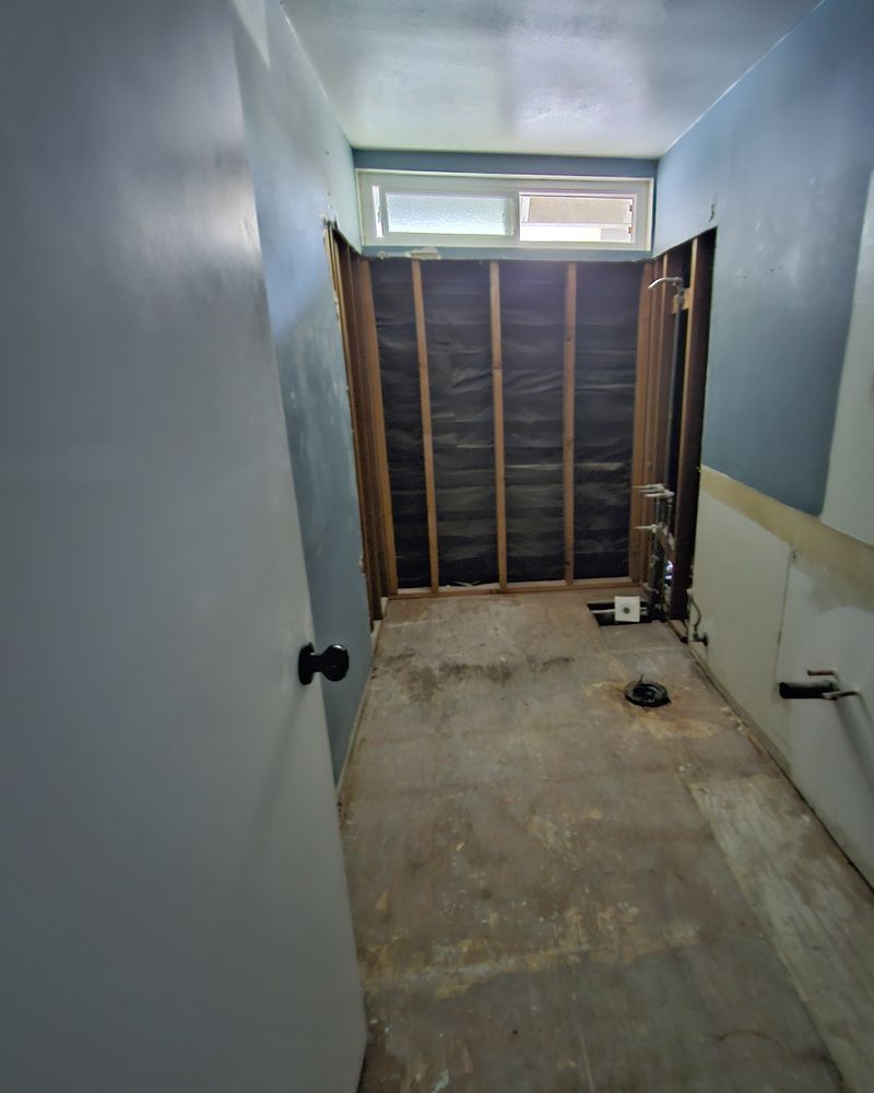 Hall bathroom gutted to the studs, with a bare subfloor and the window at the far end