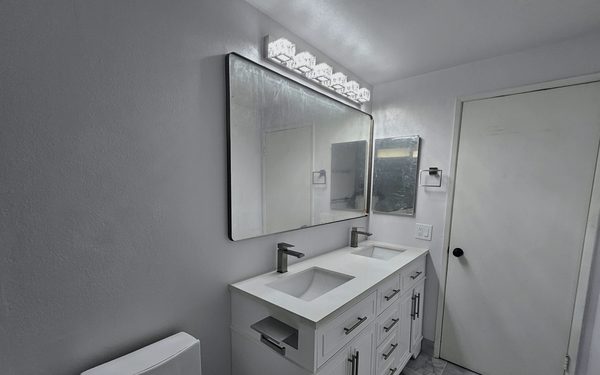 White double vanity with two faucets under a wide framed mirror and a five light bar above it