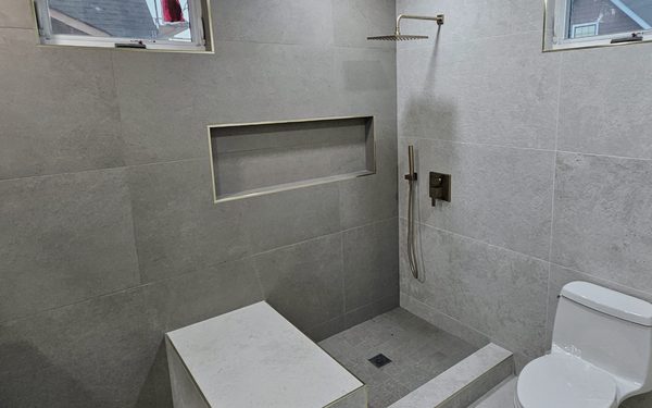 Walk in shower in large format gray tile with a built in bench, a long recessed niche, a brass rain head and a brass hand shower