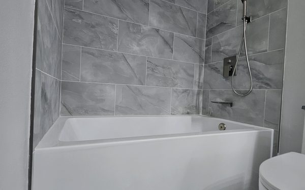 White alcove tub with a marble look gray tile surround, a black hand shower on a slide bar, and a black wall mounted valve