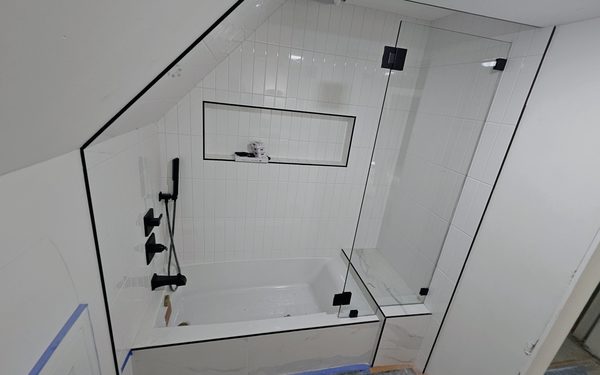 Tub set under a staircase, with white tile cut to follow the sloping ceiling, a recessed niche, black fixtures and a frameless glass panel