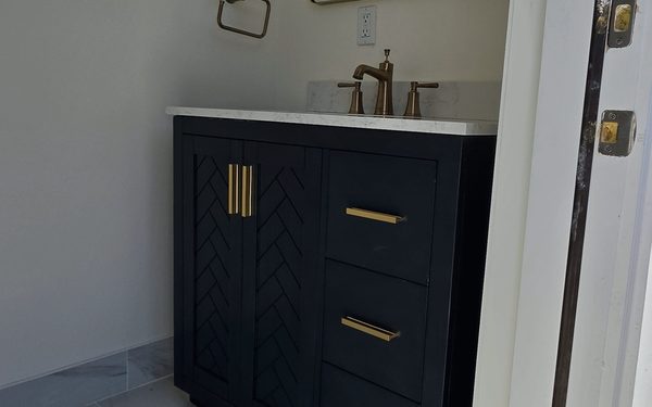 Small navy blue vanity with a herringbone cabinet front, brass pulls and a brass widespread faucet on a stone top
