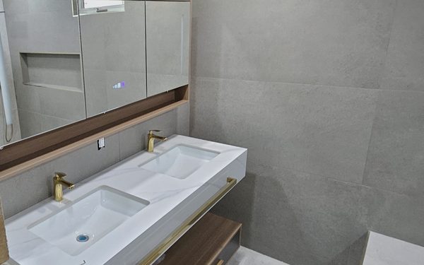Floating wood double vanity with two integrated stone basins, brass faucets, a wide mirror, and large format tile on the wall behind