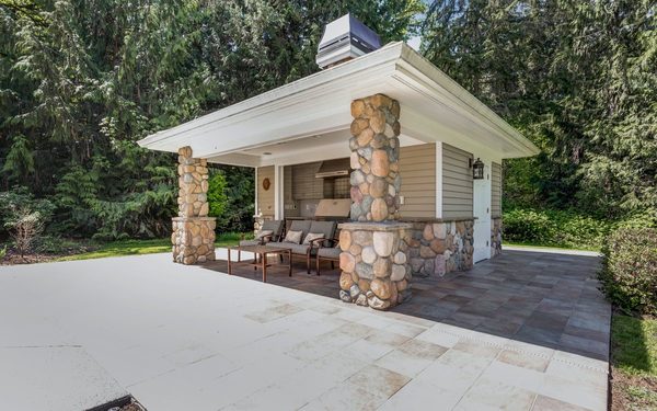 Outdoor living space with a stone clad kitchen and fireplace under a shingled roof, opening onto a paver patio
