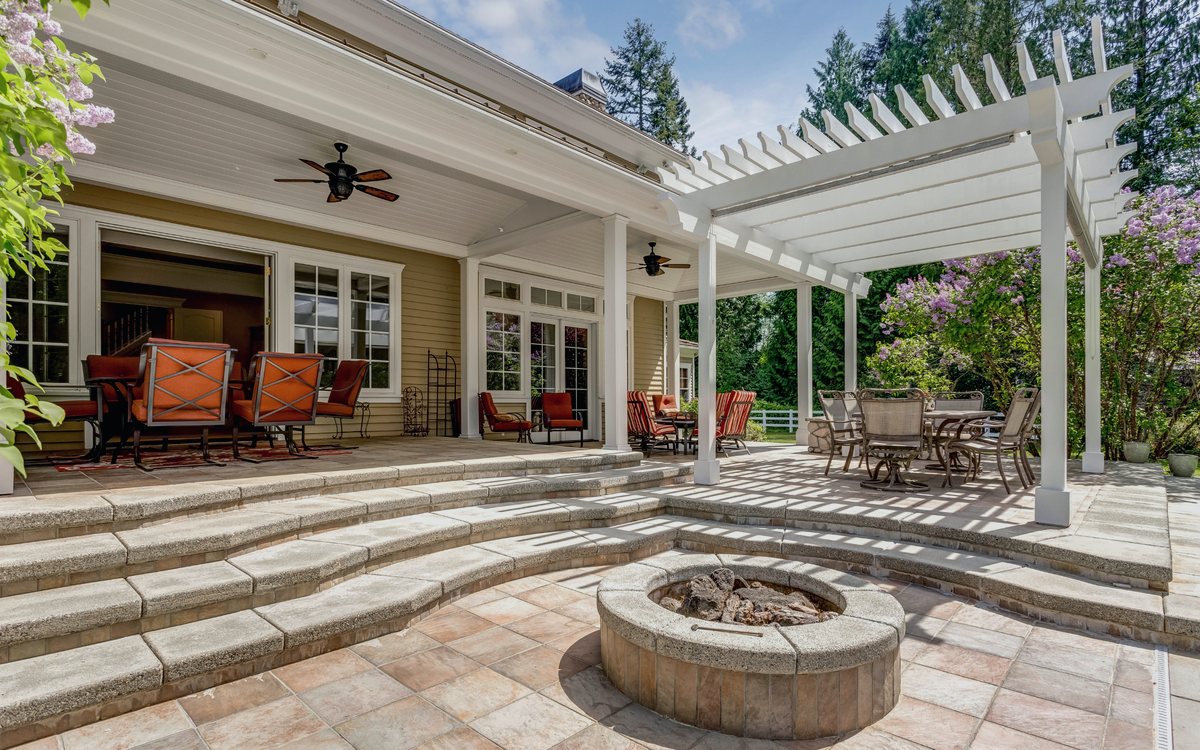 Covered patio with a white pergola, stone fire pit, and stepped paver seating area off the back of a house