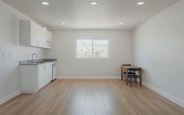 Design rendering of a plain white studio room with a short run of white kitchen cabinets and a sink along one wall, recessed ceiling lights, a single window, and a small table with two chairs