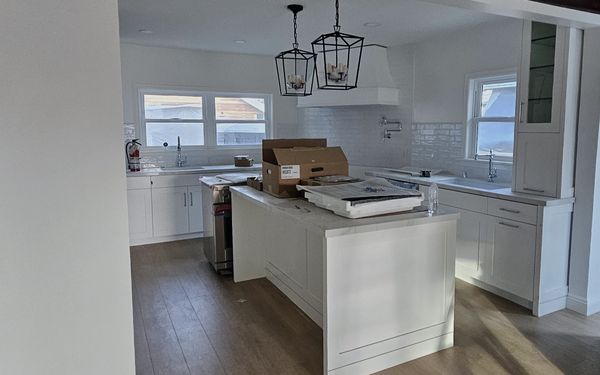 White shaker kitchen seen through a wide opening from the next room, with an island under two lantern pendants and delivery boxes still on its counter