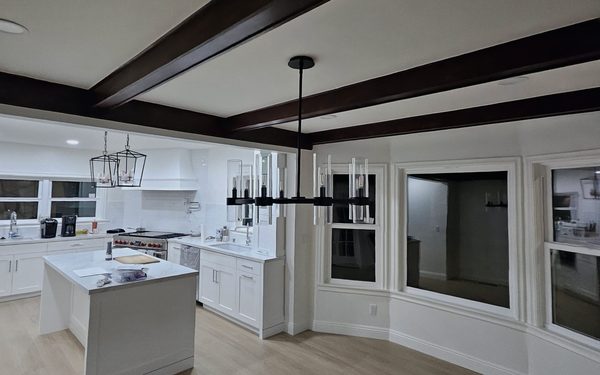 White shaker kitchen open to a dining room, with a white island under two lantern pendants and dark stained beams across the ceiling