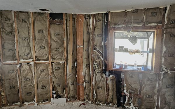 Kitchen wall opened down to the studs and insulation, with a window and copper water lines and a drain stubbed out below it
