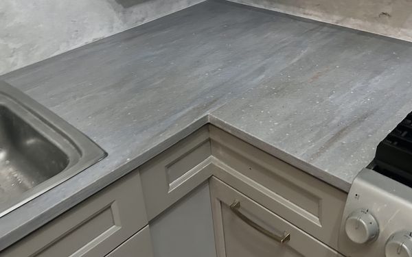 Gray counter and matching splash turning a corner between a stainless sink and a gas range, over cream cabinets with a brass pull