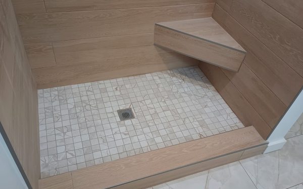 Shower tiled in wood look plank tile with a floating corner bench, a small square mosaic floor, a center drain, and metal trim closing every edge