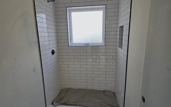 Shower alcove tiled in white subway tile around a window, with a navy patterned floor tile outside the curb