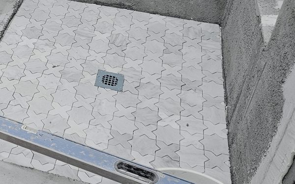 Marble star and cross mosaic being set on a shower floor around a square drain, with a level laid across the surface to check the fall
