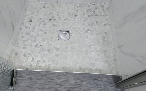 Finished hexagon marble mosaic shower floor with a square center drain, marble look wall tile, and a plank tile cap on the curb