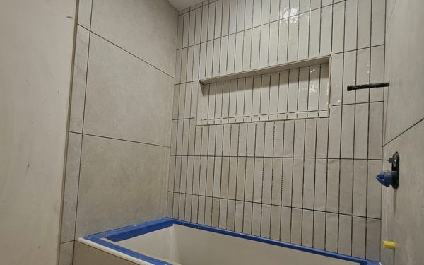 Tub alcove tiled in narrow stacked vertical tile with a long recessed niche, and large format stone look tile on the side walls